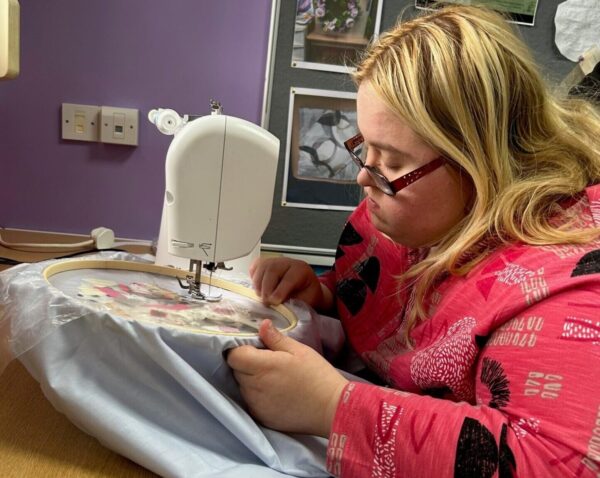 A young woman with blonde hair and glasses is focused on using a sewing machine.
